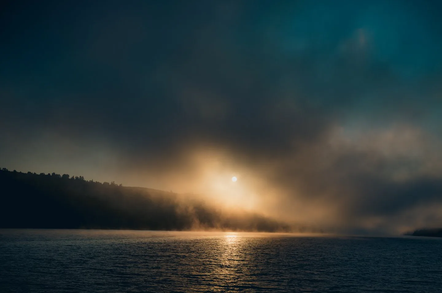 Sonnenaufgang im Nebel ueber einem See mit bewaldetem Hang in Ontario, Kanada, Lichtspur auf dem Wasser, atmosphaerische Landschaftsfotografie.