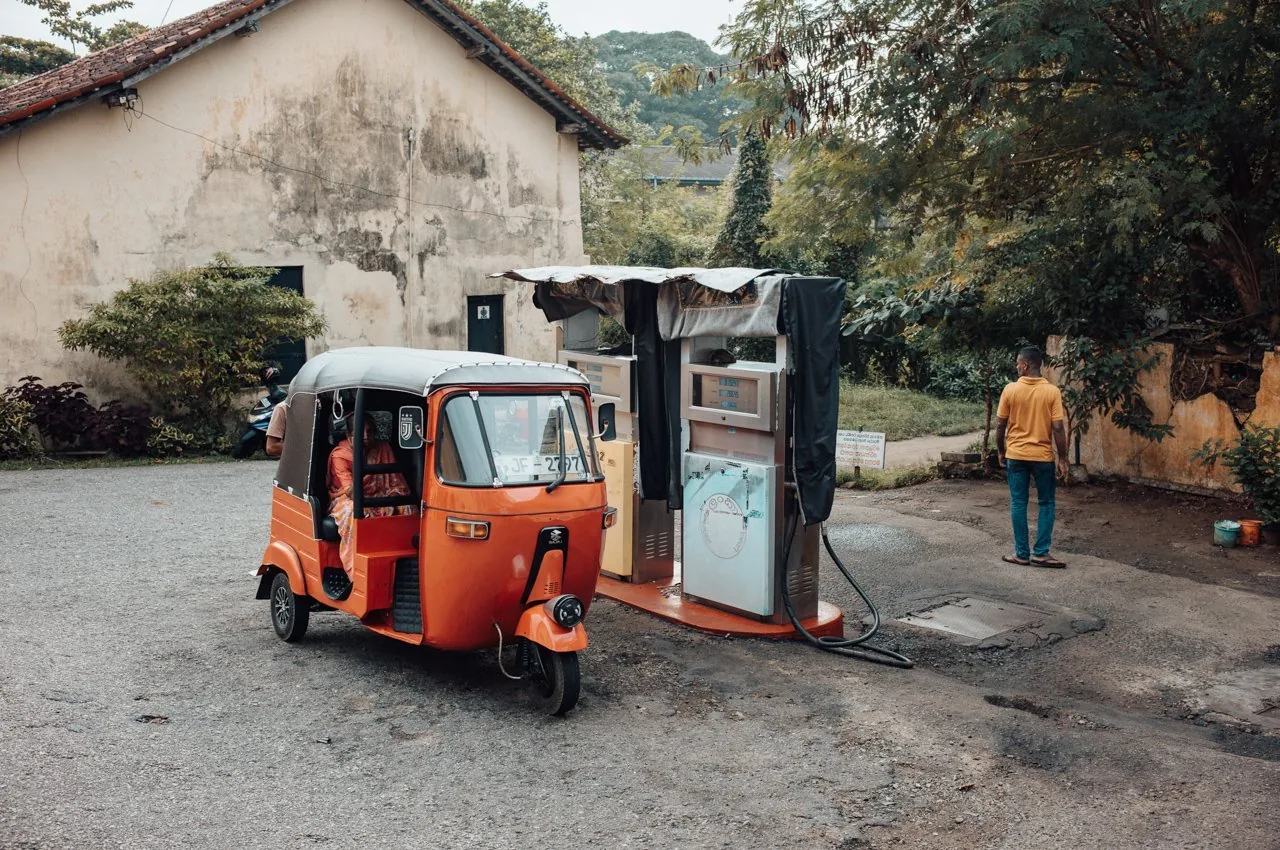 Oranges Tuk-Tuk an kleiner Tankstelle in Galle, Sri Lanka, mit verwitterter Hauswand und tropischer Vegetation.